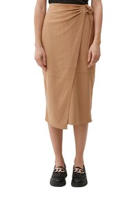Jupe en toile marron faite d'un tissu lisse avec un détail noué à la taille, présentant un ourlet diagonal. Associée à des chaussures à plateforme noires.