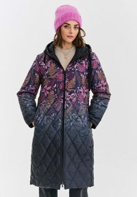 Žena oblečená v tmavém prošívaném kabátu s květinovým vzorem a předním zipem, stojící s rukama v kapsách a s jasně růžovou pletenou čepicí.