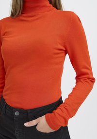 Fényes narancssárga, puha, rugalmas anyagból készült, garbós, hosszú ujjú felső. Fekete nadrág gombbal és zsebbel látható.