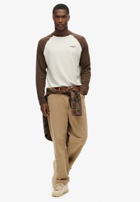 Męska koszula z długim rękawem w jasnoszarym kolorze z brązowymi rękawami raglanowymi, zestawiona z beżowymi spodniami o szerokich nogawkach i koszulą w kratę z wiązaniem w talii.