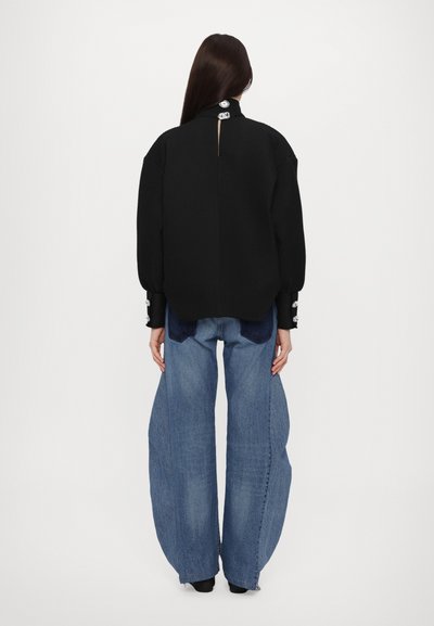 Maglione nero con texture e chiusura a bottoni sul retro, abbinato a jeans in denim blu larghi con una macchia scura.