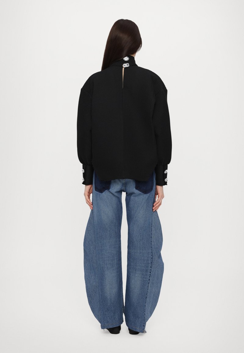 Sort tekstureret sweater med knaplukning bagpå, kombineret med løstsiddende blå denimbukser med en mørk plet.