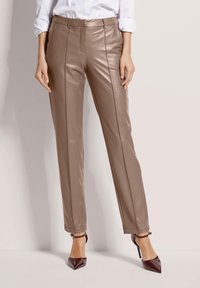 Vrouw in taupe faux lederen slim-fit broek en bordeauxrode puntige hakschoenen, staand tegen een effen achtergrond.
