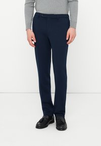 Muž v námorníckych modrých nohaviciach, čiernych kožených mokasínach s uzlíkom a svetlomodrej košeli s dlhým rukávom, stojí na bielom pozadí.