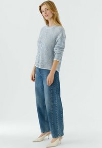 Blauw-wit gestreepte gebreide trui met lange mouwen, gecombineerd met lichtblauwe wijde jeans en crème pumps met spitse neus. Effen achtergrond.