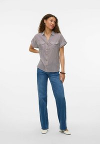 Chemise à rayures à manches courtes avec boutonnage sur le devant et deux poches poitrine ; associée à un jean bleu à jambes larges et des chaussures blanches. Fond neutre.