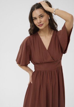 Femme aux longs cheveux bruns portant une robe marron à col en V avec des manches larges, posant avec une main sur la tête et l'autre sur la hanche.