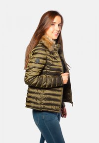 Veste matelassée vert olive avec col en fausse fourrure, fermeture éclair et poignets côtelés. Présente un patch logo sur la manche gauche.