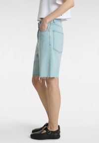 Shorts de mezclilla azul claro con dobladillo deshilachado, diseño de cinco bolsillos y costuras visibles. Combinados con zapatos negros con tachuelas decorativas.