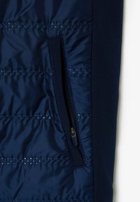 Tela acolchada azul marino con costuras horizontales y un bolsillo vertical con cremallera en el lado derecho, mostrando el detalle del tirador de la cremallera.