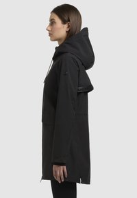 Black waterproof jacket with a hood, featuring a front zip, side pockets, and a logo-accented strap on the shoulder. Textured fabric.