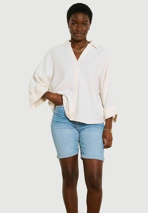Vrouw die een los wit overhemd met knopen en lichtblauwe denim shorts draagt, staand met één hand in haar zak tegen een effen achtergrond.
