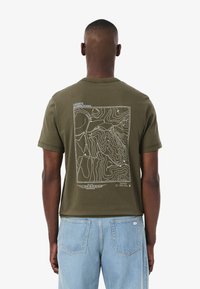 Camiseta de algodón verde oliva con un gráfico de un mapa topográfico blanco en la parte posterior, mangas cortas y cuello redondo.