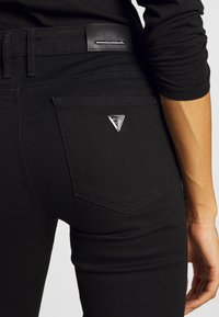 Černé džínové kalhoty se trojúhelníkovým kovovým logem na zadní kapse, slim fit a hladkým povrchem s viditelným prošitím.