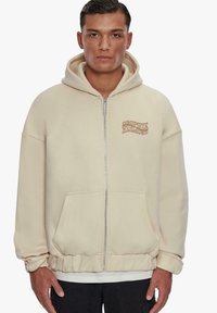 Beige zip-up hoodie made of soft fabric with a kangaroo pocket, ribbed hem, and contrasting logo on left chest. Adjustable drawstring hood.