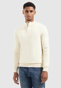 Man die tegen een effen achtergrond staat, gekleed in een crèmekleurige ritstrui en blauwe jeans, met een neutrale uitdrukking naar voren gericht.