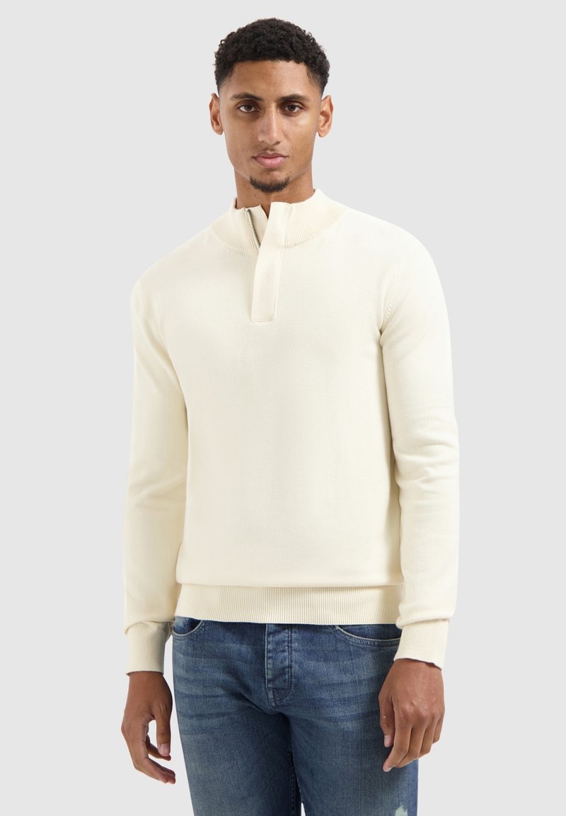 Man die tegen een effen achtergrond staat, gekleed in een crèmekleurige ritstrui en blauwe jeans, met een neutrale uitdrukking naar voren gericht.