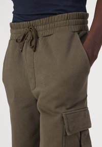 Olive green jogger pants with an elastic waistband, drawstring closure, side pockets, and a cargo pocket on the thigh. Soft fabric texture.