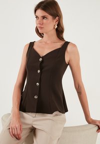 Rjava brezrokavna majica z V-izrezom in petimi srebrnimi gumbi na sprednji strani. Gladek material z oprijetim pasom in razširjenim robom.