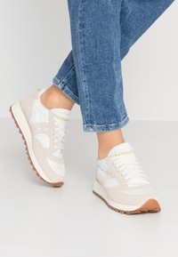 Baskets avec une tige en suède beige et en textile blanc brillant, accentuées par un logo doré, des lacets blancs et une semelle en caoutchouc marron clair.