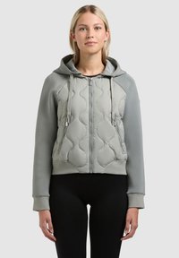 Mladá žena v světle šedé prošívané mikině na zip se šňůrkami a černých kalhotách, stojící před jednobarevným pozadím.