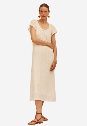 Knielanges, beiges Strickkleid mit kurzen Ärmeln, strukturiertem Muster und rundem Ausschnitt. Kombiniert mit braunen Sandalen mit Riemchen.