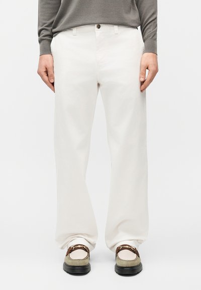 Personne portant un pantalon blanc ample, des mocassins beige et marron, et un haut gris à manches longues, debout devant un fond uni.