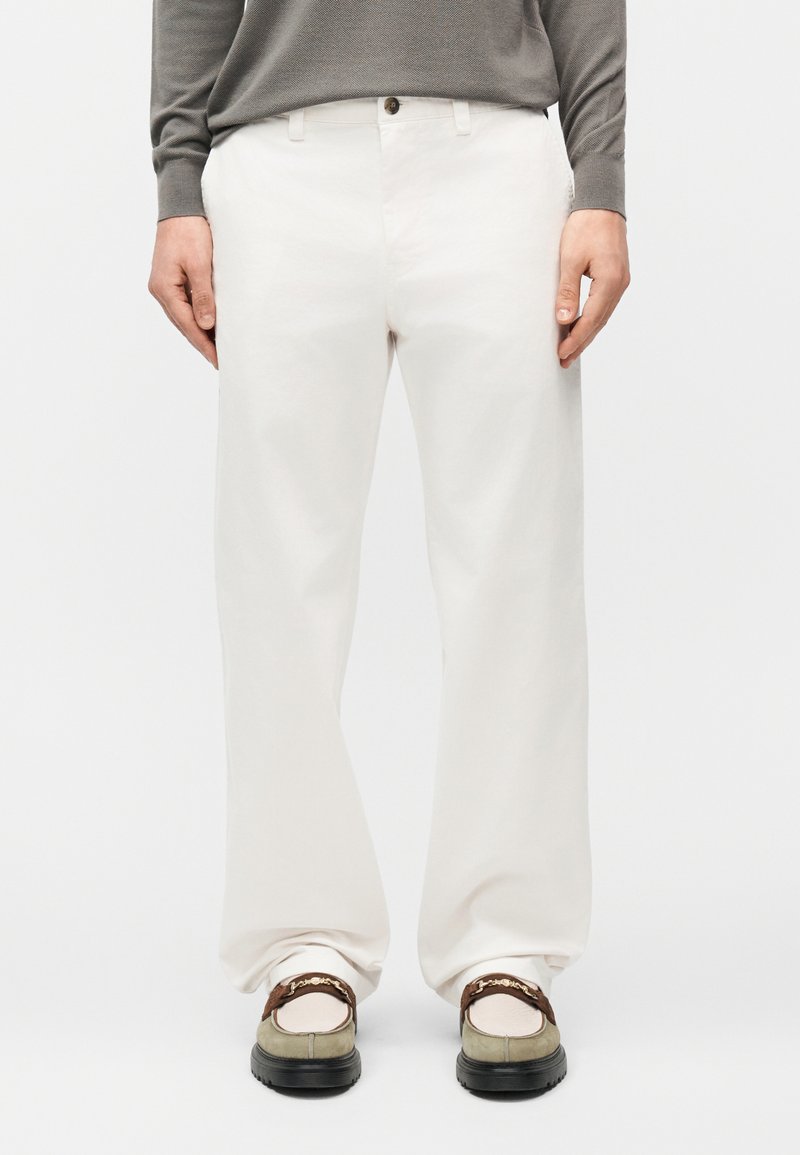 Personne portant un pantalon blanc ample, des mocassins beige et marron, et un haut gris à manches longues, debout devant un fond uni.