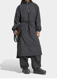 Mustan kevyt vedenpitävä takki, jossa on korkea kaulus ja kiristettävä vyötärö, yhdistettynä löysiin mustiin housuihin ja puusandaaleihin. Yksinkertainen muotoilu.