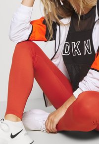 Leggings rossi abbinati a una maglietta in rete nera e scarpe da ginnastica bianche e testurizzate. La giacca presenta dettagli arancioni e una chiusura con zip.