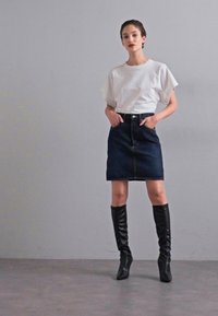 Biela blúzka s krátkym rukávom v kombinácii s tmavými džínovými sukňami. Modelka má na sebe čierne vysoké čižmy po kolená a stojí na neutrálnom pozadí.