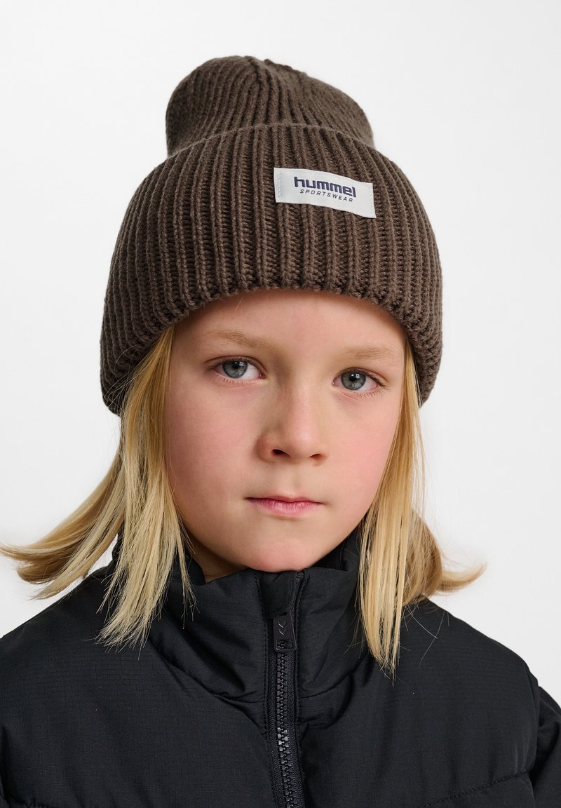 Bruine geribbelde gebreide beanie met een omgeslagen rand en een wit label met de tekst "hummel sportswear," gedragen door een kind in een zwarte ritsjack.