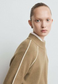 Persona joven con el cabello peinado hacia atrás que lleva un suéter de punto beige con rayas blancas, mirando hacia la cámara sobre un fondo liso.