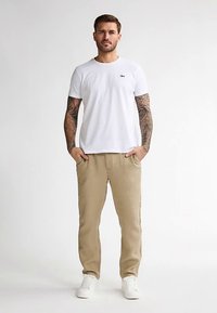 Witte T-shirt met ronde hals, beige broek met zijzakken en witte sneakers. Heeft tatoeages op de armen, staat tegen een neutrale achtergrond.