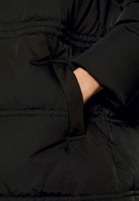 Schwarze Daunenjacke mit glatter, glänzender Oberfläche, gestepptem Design und einer seitlichen Tasche, aus der eine Hand teilweise sichtbar ist.