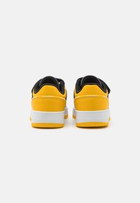 Svart og gul sneakers med glatt overflate, polstrede krager, hvite mellomsåler og teksturerte gummisåler. Velcro-stroppdetalj på sidene.