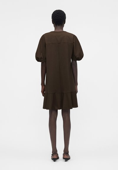 Άτομο στέκεται με την πλάτη προς τα εμπρός, φοράει ένα φαρδύ σκούρο καφέ φόρεμα με φουσκωτά μανίκια και μαύρα σανδάλια με τακούνι σε απλό φόντο.
