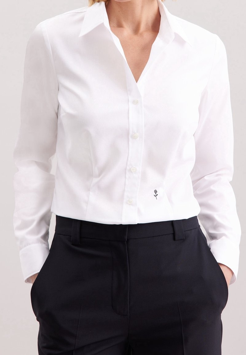 Femme portant une chemise blanche à boutons avec un petit emblème de fleur noire près de la taille et un pantalon noir sur mesure, les mains dans les poches.