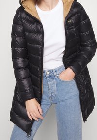 Sort pufferjakke med hætte, med en tan-farvet indvendig foring, lynlås lukning og quiltet tekstur. Båret over en hvid skjorte og blå jeans.