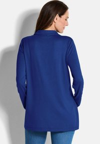 Langärmliges Oberteil in einfarbigem Blau mit einer weichen Textur und einem Kragen. Das Design ist lässig geschnitten und endet auf Hüfthöhe, kombiniert mit blauen Jeans.