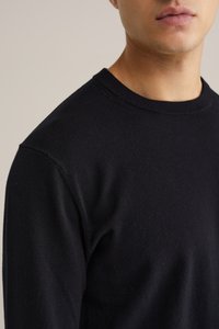 Melnā adītā džemperī ar teksturētu virsmu, kas piedāvā apaļu kakla izgriezumu un ribotas aproces. Tuvplāns skats uz audumu un kakla izgriezuma detaļām.