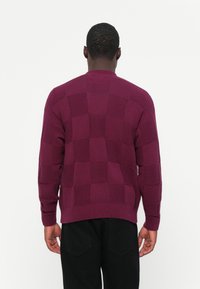 Maglione knit bordeaux con un motivo a scacchi, colletto e polsini a coste, che mette in risalto una superficie testurizzata. Indossato al contrario, rivelando dei dettagli.
