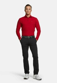 Rotes Langarm-Polohemd mit Kragen, schwarze Hose und einen mehrfarbigen gemusterten Gürtel. Die Schuhe sind hellgrau mit weißen Akzenten.