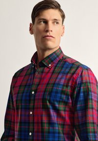 Camisa de cuadro de hombre con botones que presenta colores rojo, verde y azul, con un cuello en contraste y un pequeño logo en el pecho. La textura parece suave.