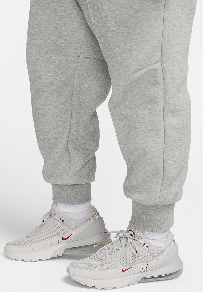 Helle graue Sweatpants mit gerippten Bündchen, kombiniert mit weißen Sneakers, die eine strukturierte Oberseite, rote Akzente und eine sichtbare Luftpolsterung aufweisen.