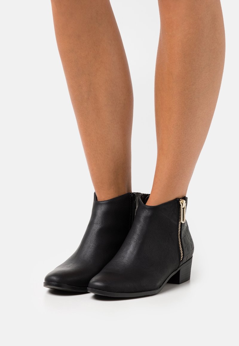 Schwarzlederne Stiefeletten bis Knöchelhöhe mit seitlichen Reißverschlüssen, die auf nackten Beinen getragen werden. Sie haben einen niedrigen Blockabsatz und eine leicht abgerundete Zehenpartie.