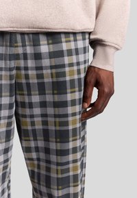 Pantalon à carreaux gris et olive avec une finition lisse, assorti d'un sweat-shirt beige clair. Comprend une main visible reposant sur le pantalon.