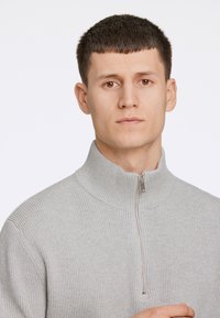 Ljusgrå räfflad tröja med hög krage och halvdragkedja vid halsen, som har en slät textur och en figurnära design.