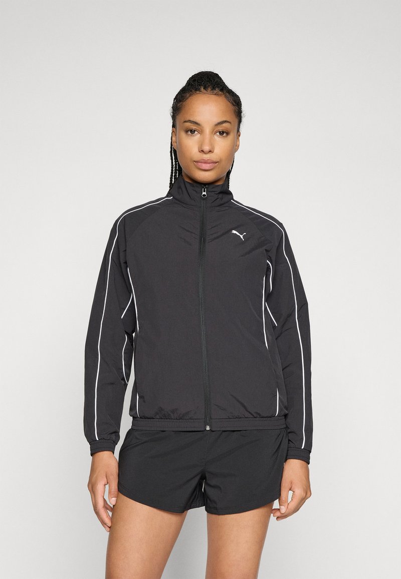 Mulher de pé, de frente, vestindo uma jaqueta preta da Puma com zíper e detalhes brancos, juntamente com calções desportivos pretos, contra um fundo simples.