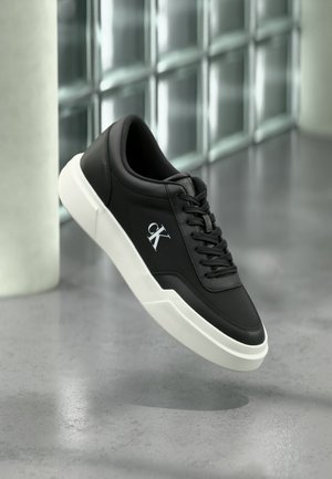 Μαύρο δερμάτινο αθλητικό παπούτσι με λευκή σόλα και λογότυπο "cK", εμφανίζεται στον αέρα μπροστά από τσιμεντένιο δάπεδο και τοίχο από γυάλινους κύβους.